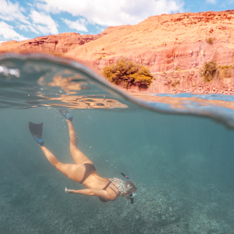 Snorkeler with fins swims over a coral reef in a split-view image with Molokini’s rocky crater wall above the water