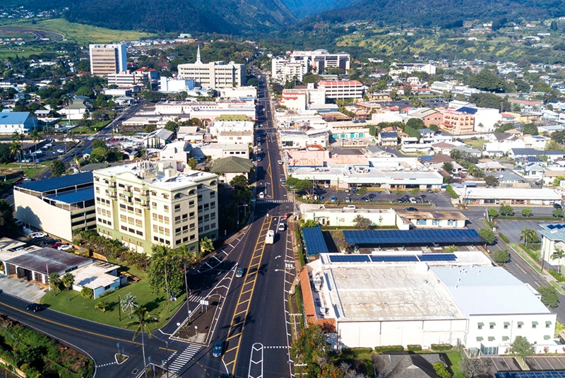 Aerial view of Wailuku, Maui with town buildings, roads, parked cars, and green mountains rising in the distance.