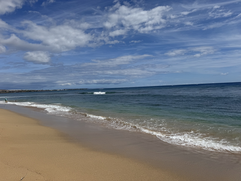 La playa de Kaanapali en Maui, con su costa arenosa, las suaves olas del océano y un cielo azul nublado.