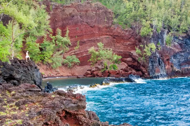 Kaihalulu Red Sand Beach in Hana Maui with red cliffs volcanic rock shoreline and blue ocean water.