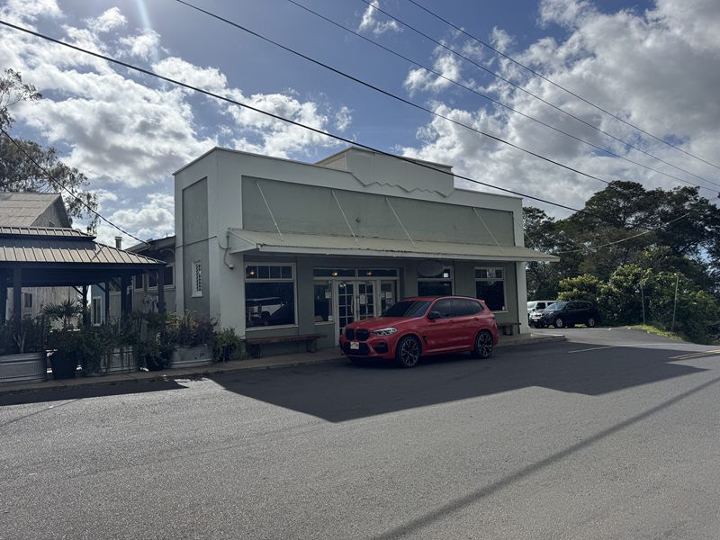 Außenansicht des Kula Bistro auf Maui mit Parkplätzen an der Straße, Pflanzen im Außenbereich und einem davor geparkten roten SUV.