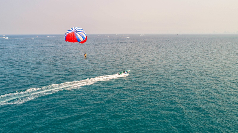 Un practicante de parapente flota a gran altura sobre el océano abierto mientras una pequeña embarcación surca el agua a toda velocidad en un día despejado.