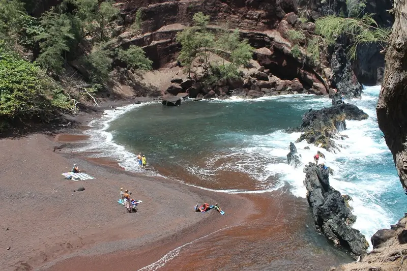 Red Sand Beach in Hana Maui with red shoreline volcanic cliffs ocean waves and beachgoers relaxing.