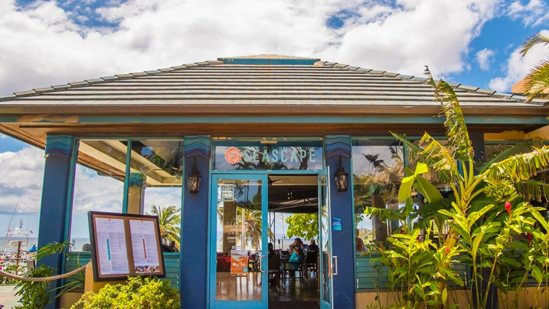 Entrance to Seascape Restaurant with tropical plants, posted menus, blue trim, and Maʻalaea Harbor boats nearby