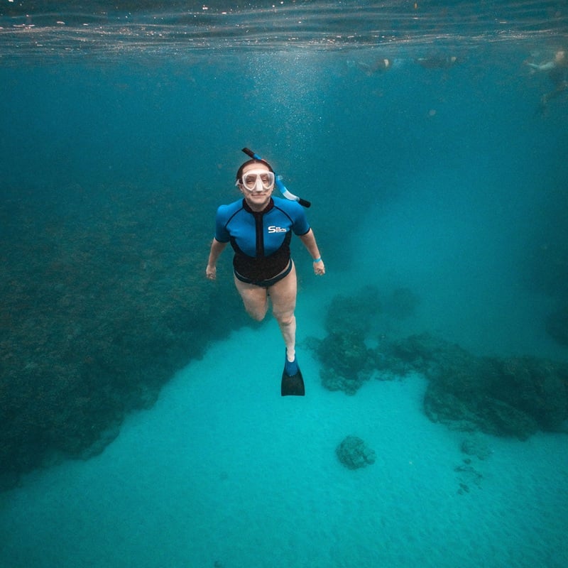 Snorkeler wearing a mask, snorkel, blue flotation top, and visible fin floats underwater in clear Maui water.
