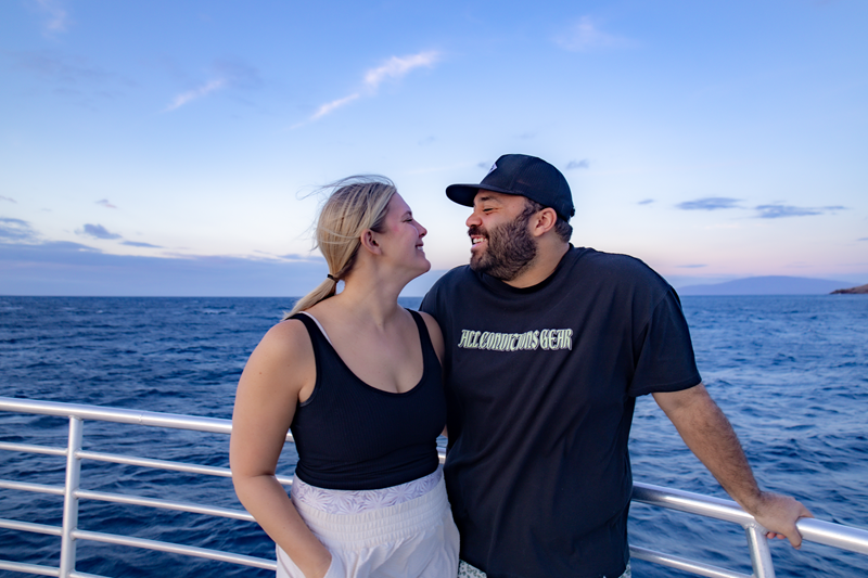 Two guests smiling together on a Maui boat tour with ocean, coastline, and a soft evening sky in the background