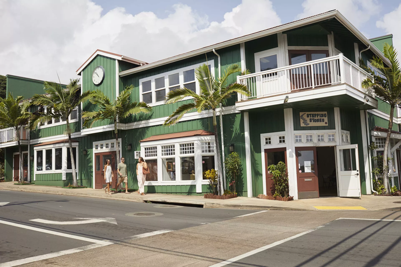 Green storefronts along a Wailuku, Maui street with palm trees, sidewalks, and people walking past local shops.